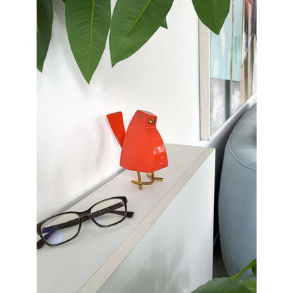 Moderne geometrische Kunstharzskulptur: Kleiner Vogel in leuchtendem Orange, stylisch platziert auf einem Regal neben einer Brille, umgeben von Pflanzen.