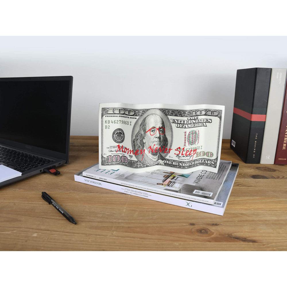 Pop Art Tischskulptur "Money Never Sleeps" - Tischszene mit einer 100-Dollar-Banknote, die als künstlerisches Objekt betrachtet wird, umgeben von einem Laptop und Zeitschriften.